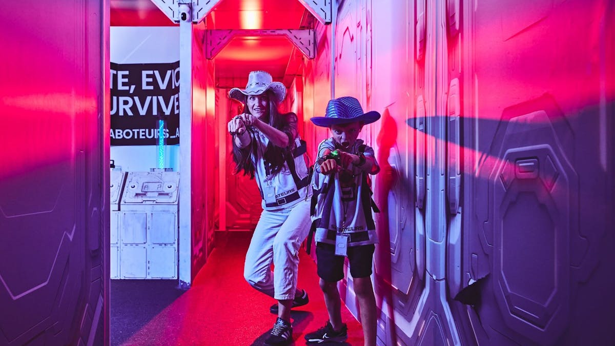 Mother and son in Saboteurs gear and cowboy hats creep through a red-lit corridor.