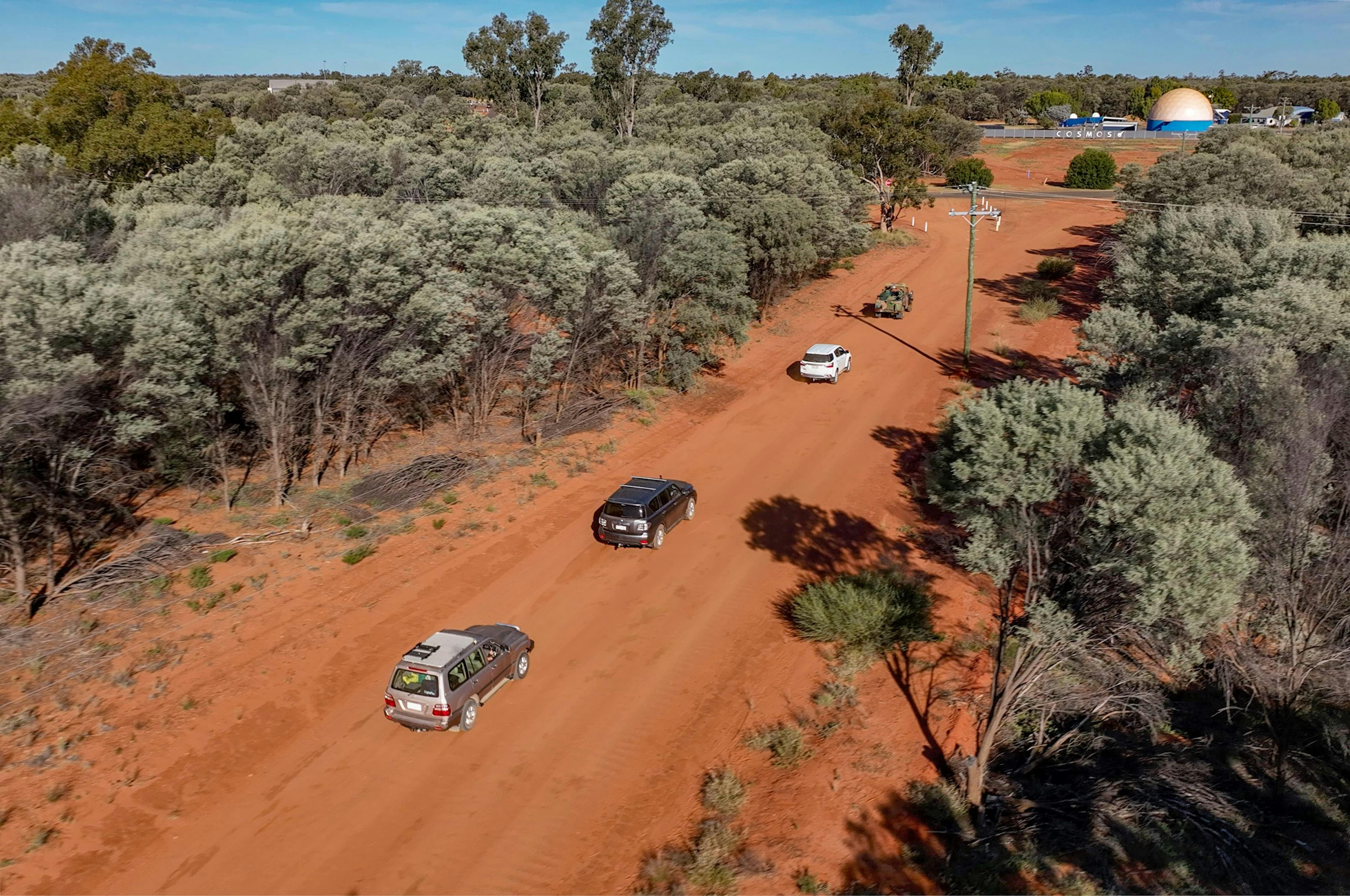 aerial shot of cars travelling in a convoy on a dirt road surrounded by Mulga trees