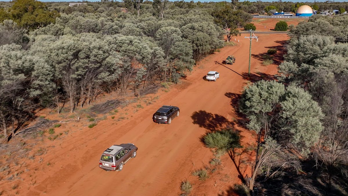 aerial shot of cars travelling in a convoy on a dirt road surrounded by Mulga trees