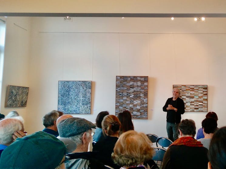An artist speaks to a seated audience with his artworks on the wall behind him.