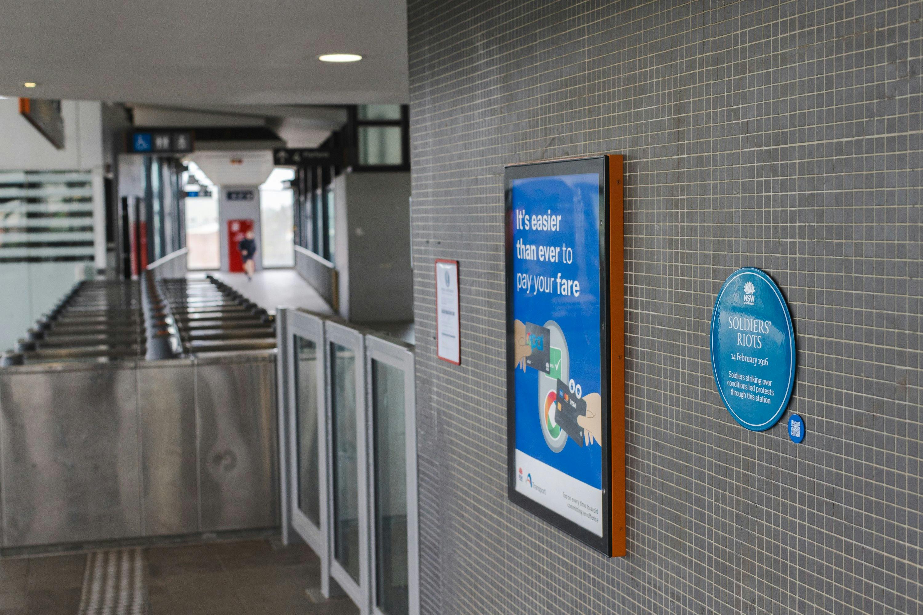 Blue plaque on tiled grey wall next to a poster and turnstiles in background