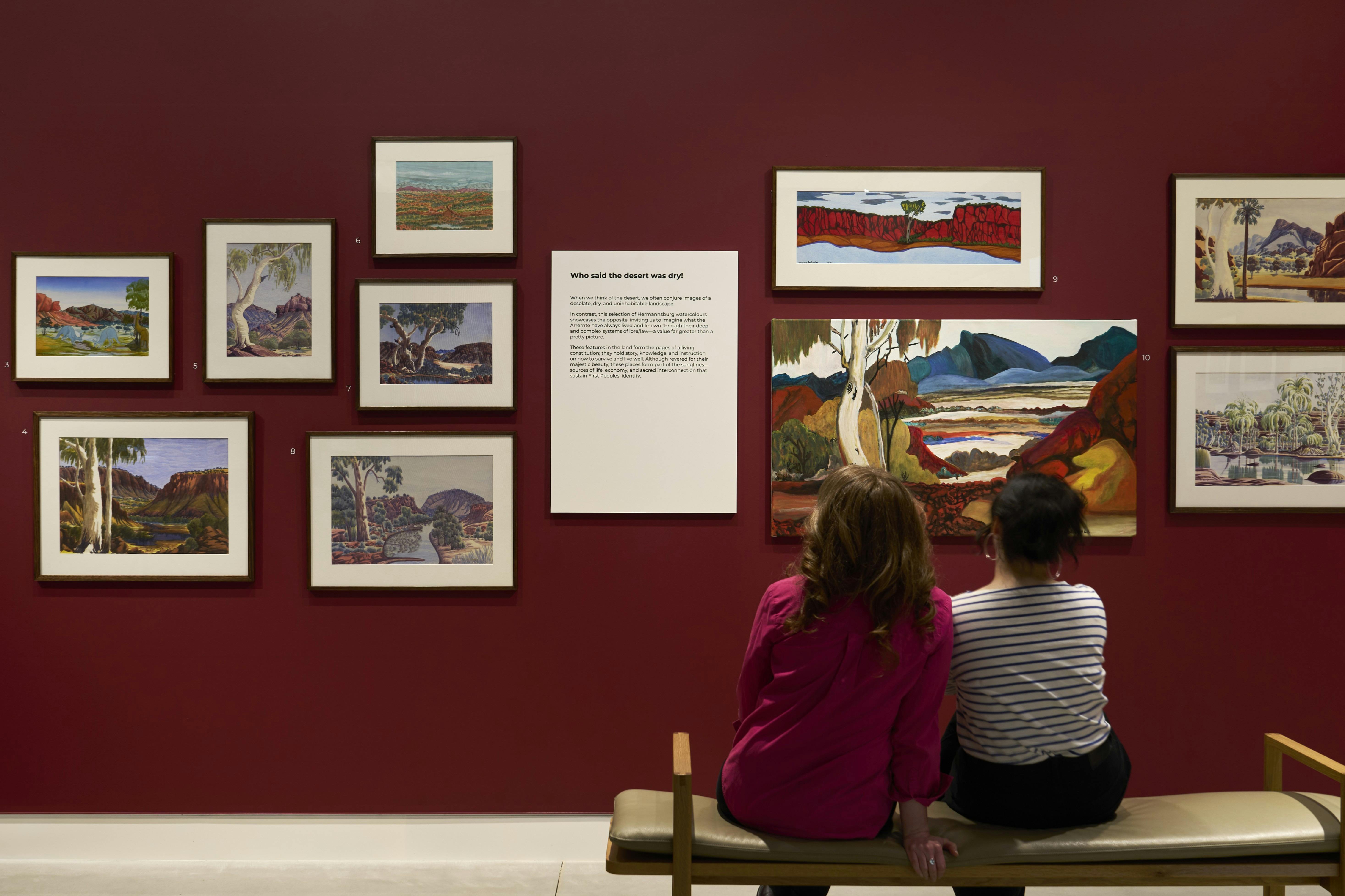 Two women sit on a bench in front of a wall of paintings by Indigenous artists arranged salon-style