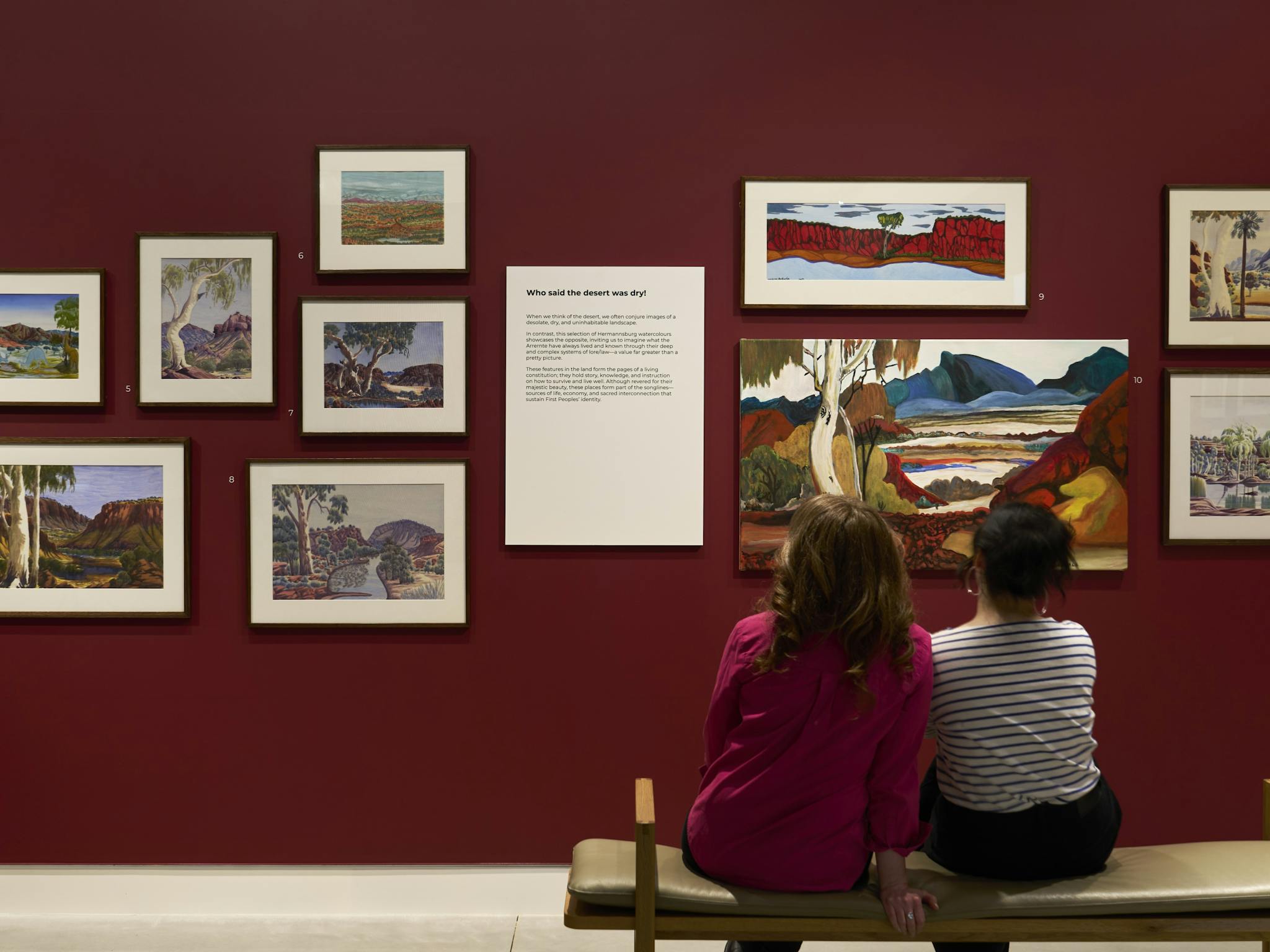 Two women sit on a bench in front of a wall of paintings by Indigenous artists arranged salon-style