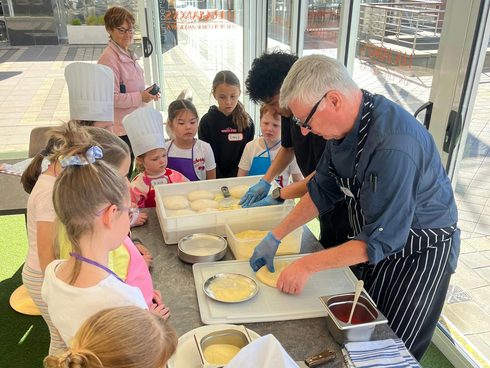 Two Chefs bent over a table showing children how to stretch out pizza dough