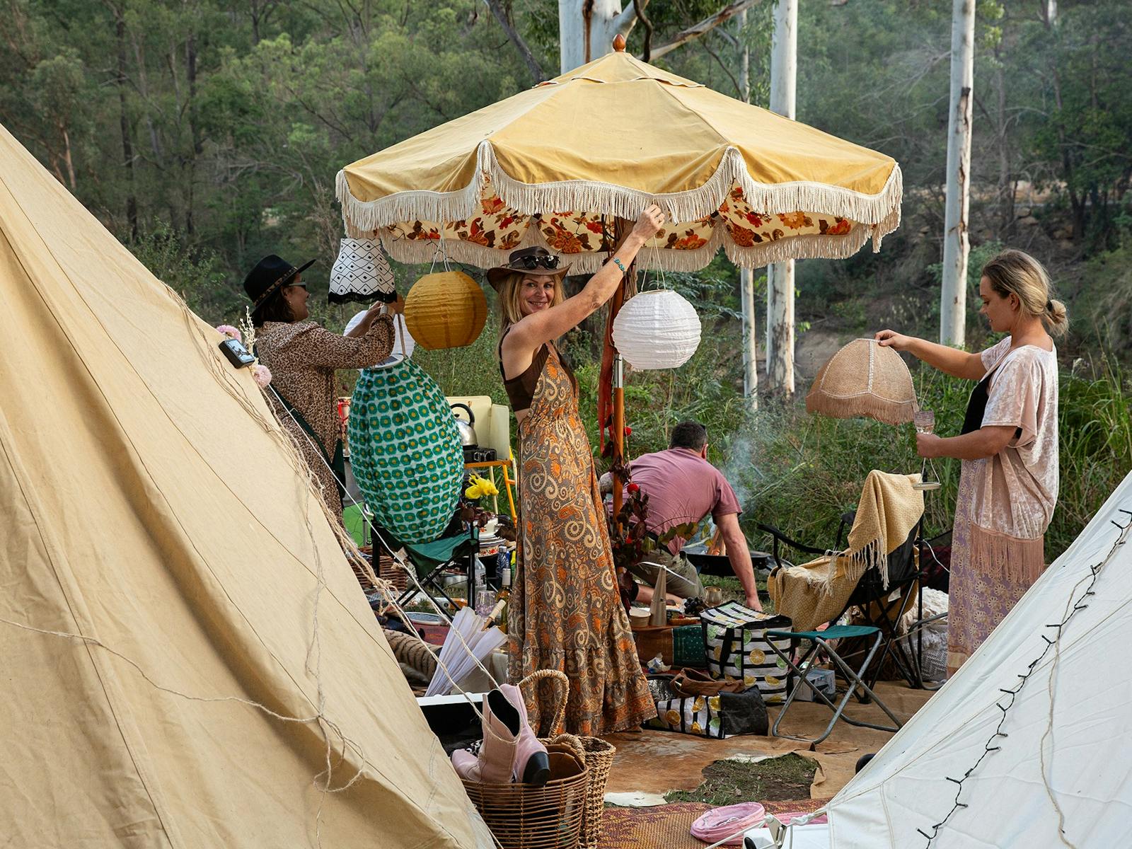 Women in cotton dresses hang paper lamps from a beach umbrella in a campsite