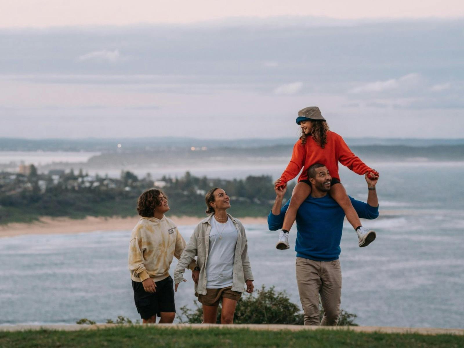 Two adults, and a boy walking with a young girl on a man shoulders, with ocean in the background