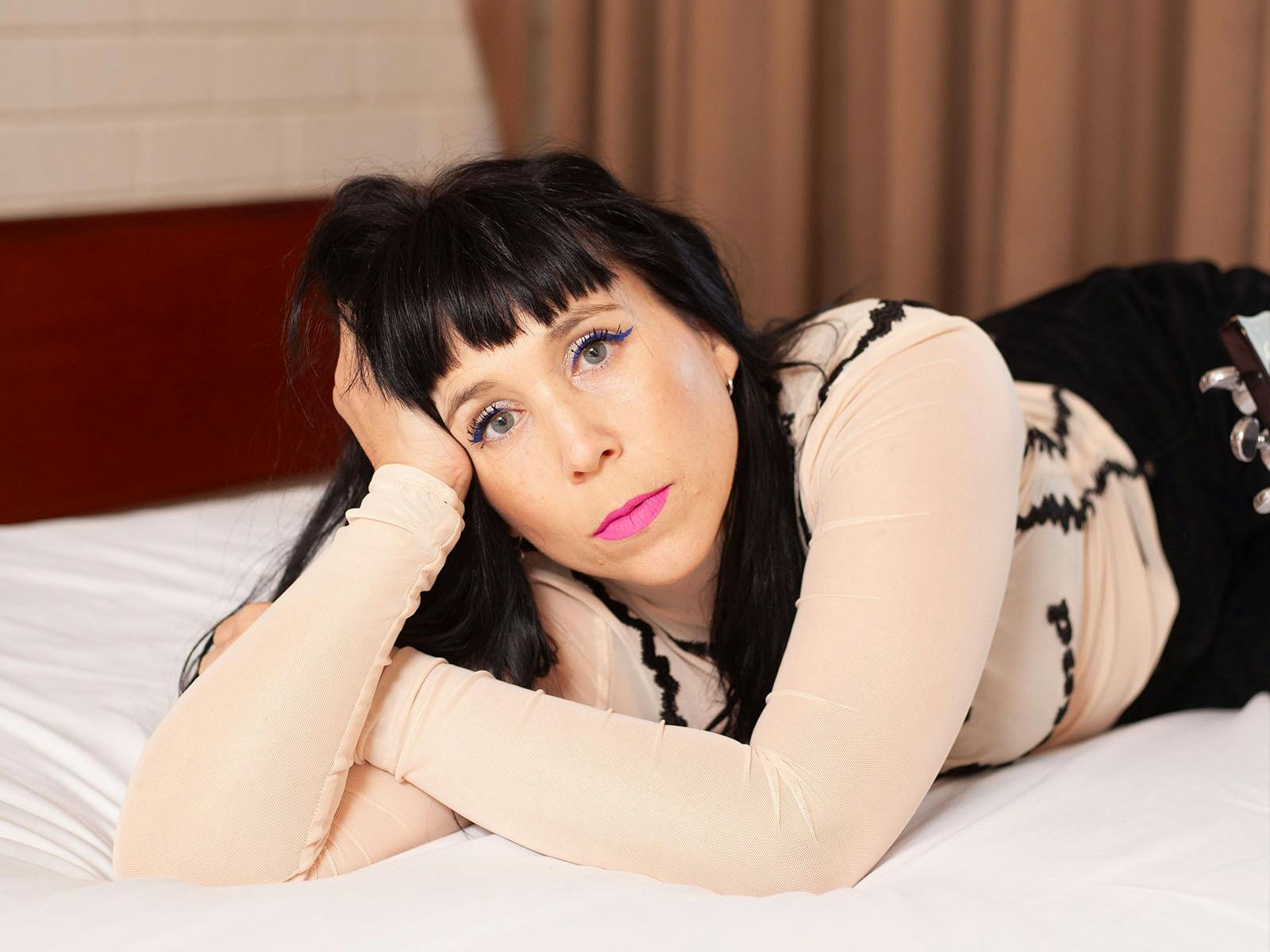 A white woman with pink lipstick wearing a taupe shirt and black pants, rests on her elbow on a bed