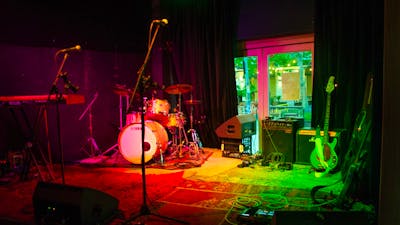Image of a drumset, microphone, a couple of amps on a stage with red and green lighting.