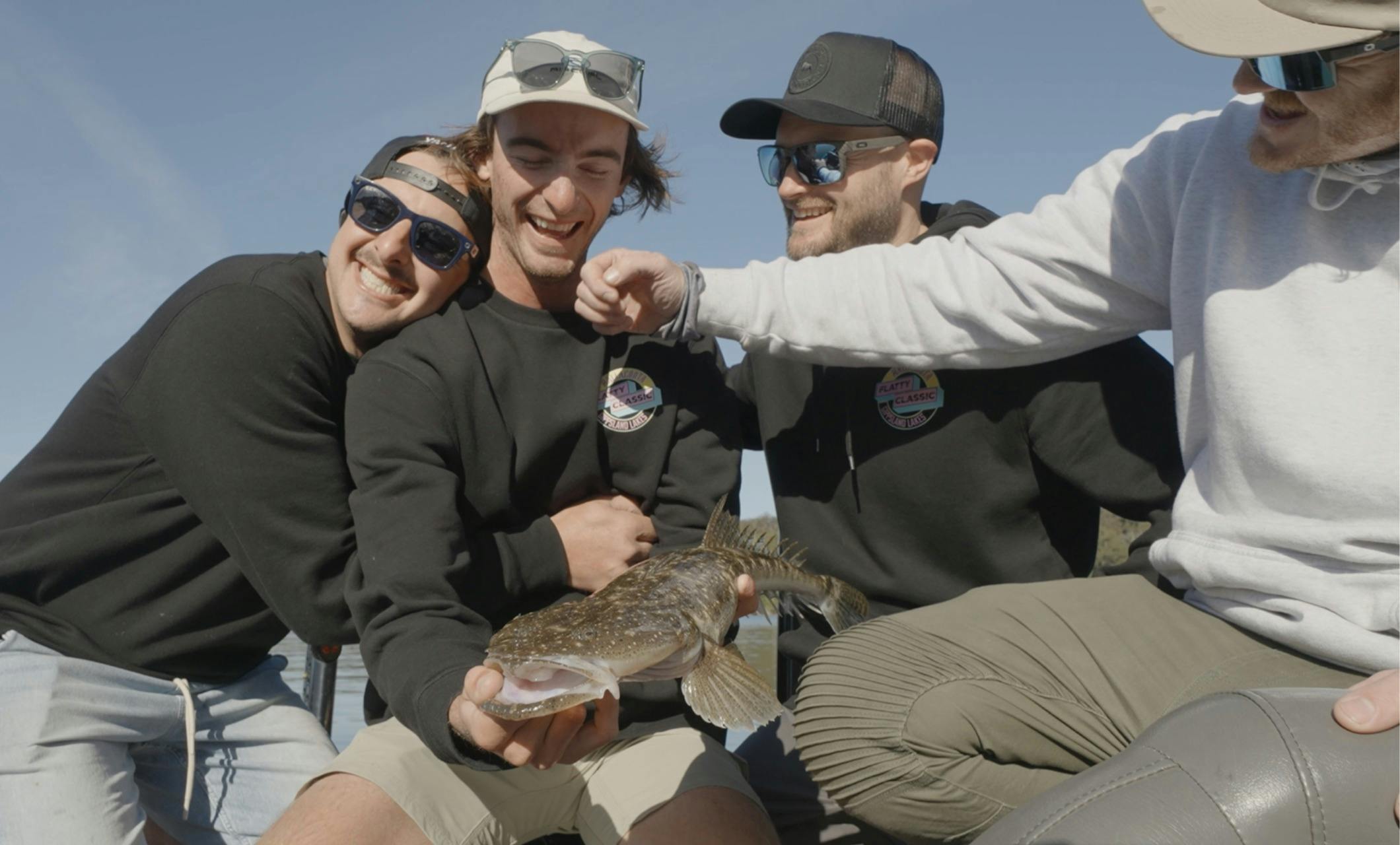 Four smiling teammates in Flatty Classic jumpers crowd around a dusky flathead
