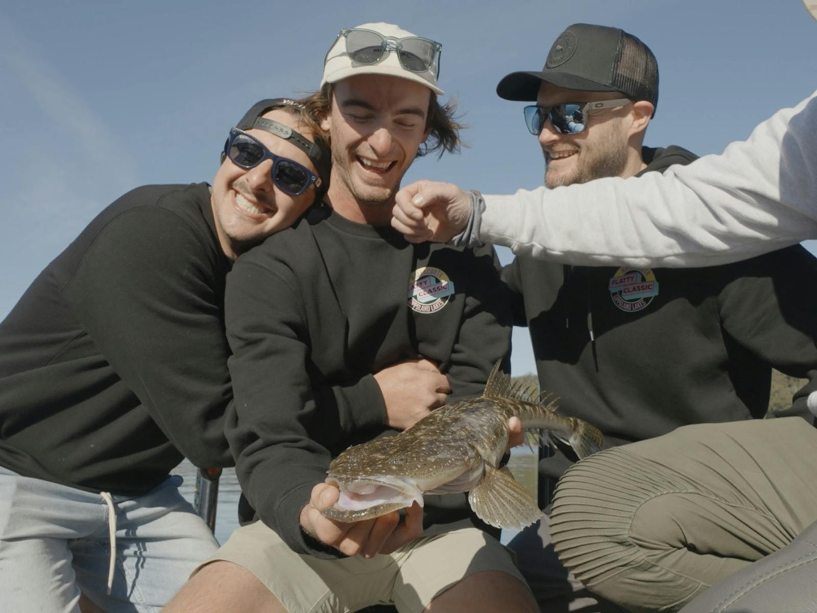 Four smiling teammates in Flatty Classic jumpers crowd around a dusky flathead