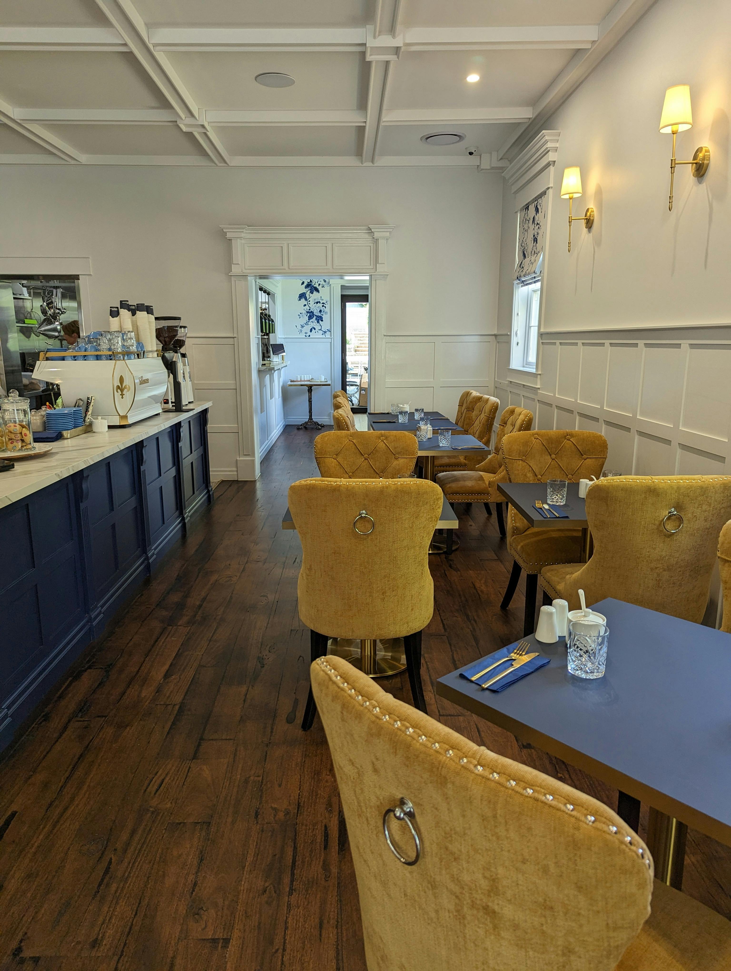Dark timber tables and cushioned yellow dining chairs in the cafes front room. The counter is blue .