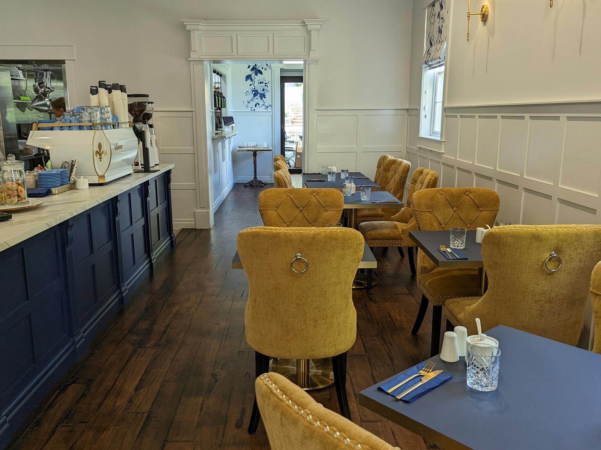 Dark timber tables and cushioned yellow dining chairs in the cafes front room. The counter is blue .
