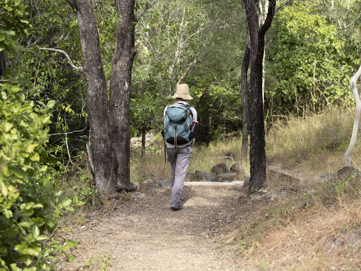 Person walking along track in long pants, shirt and hat, carrying a backpack.