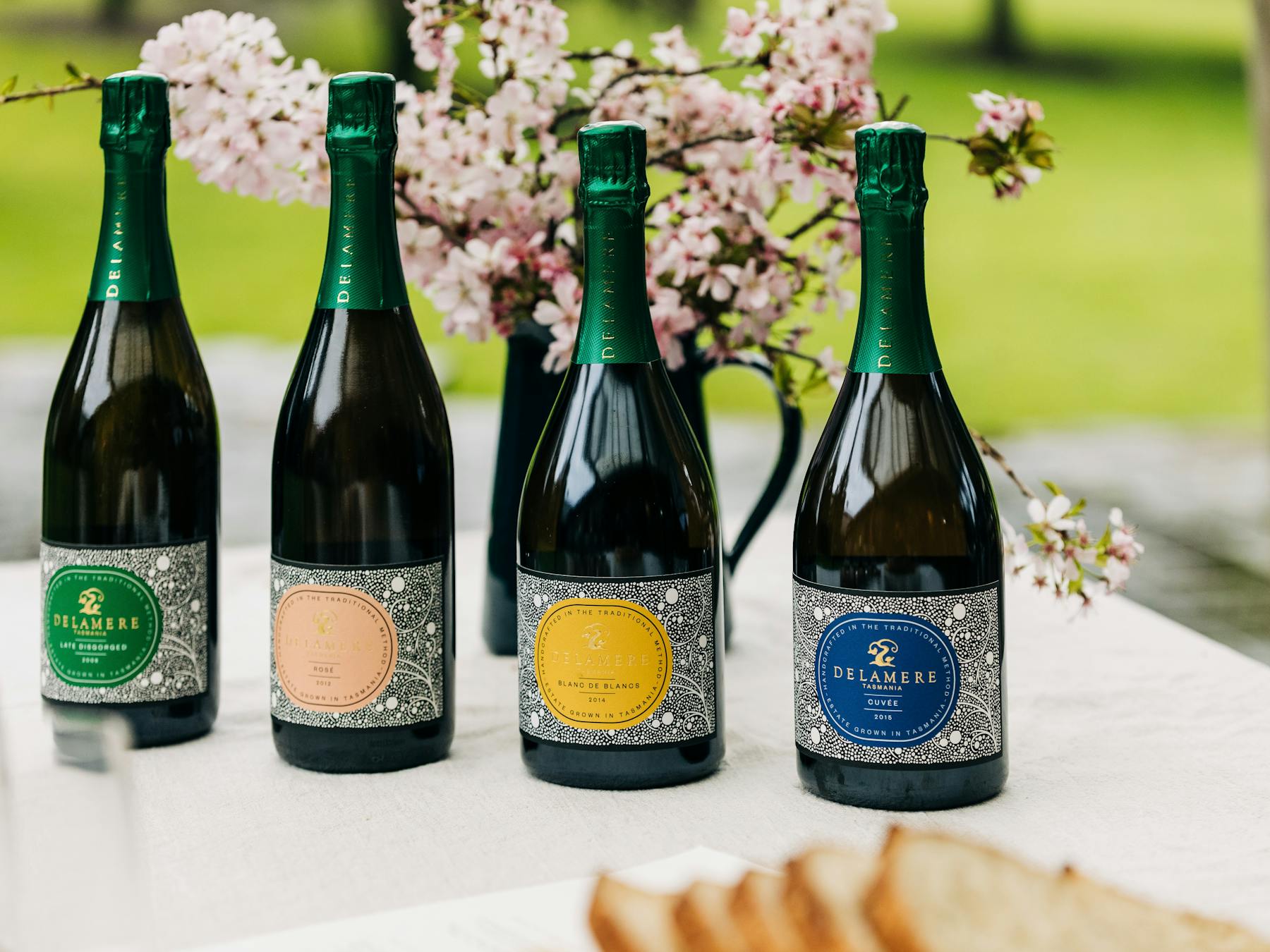 Four bottles of Delamere sparkling wine on picnic table with cherry blossom in the background.