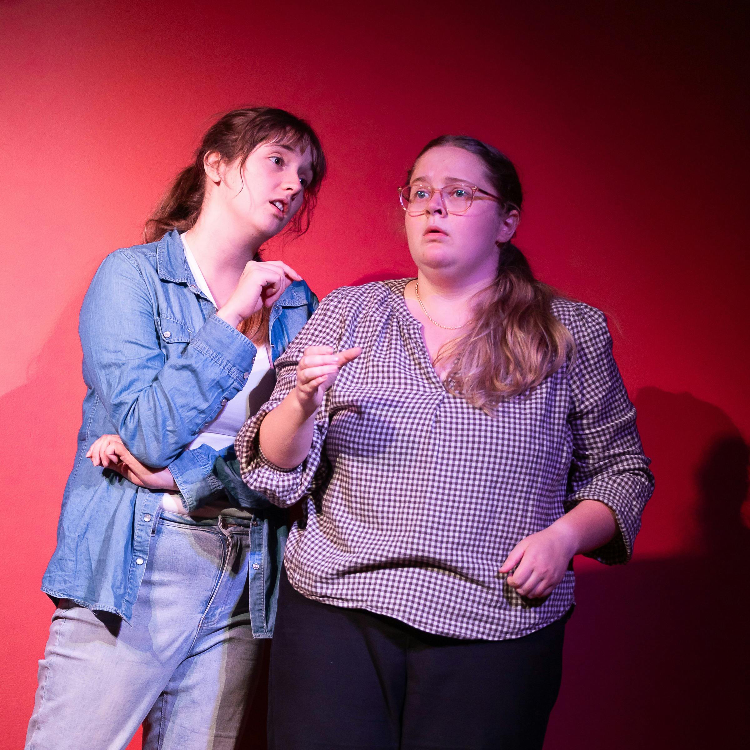 Two young women stand close together. One goads the other, who appears anxious.