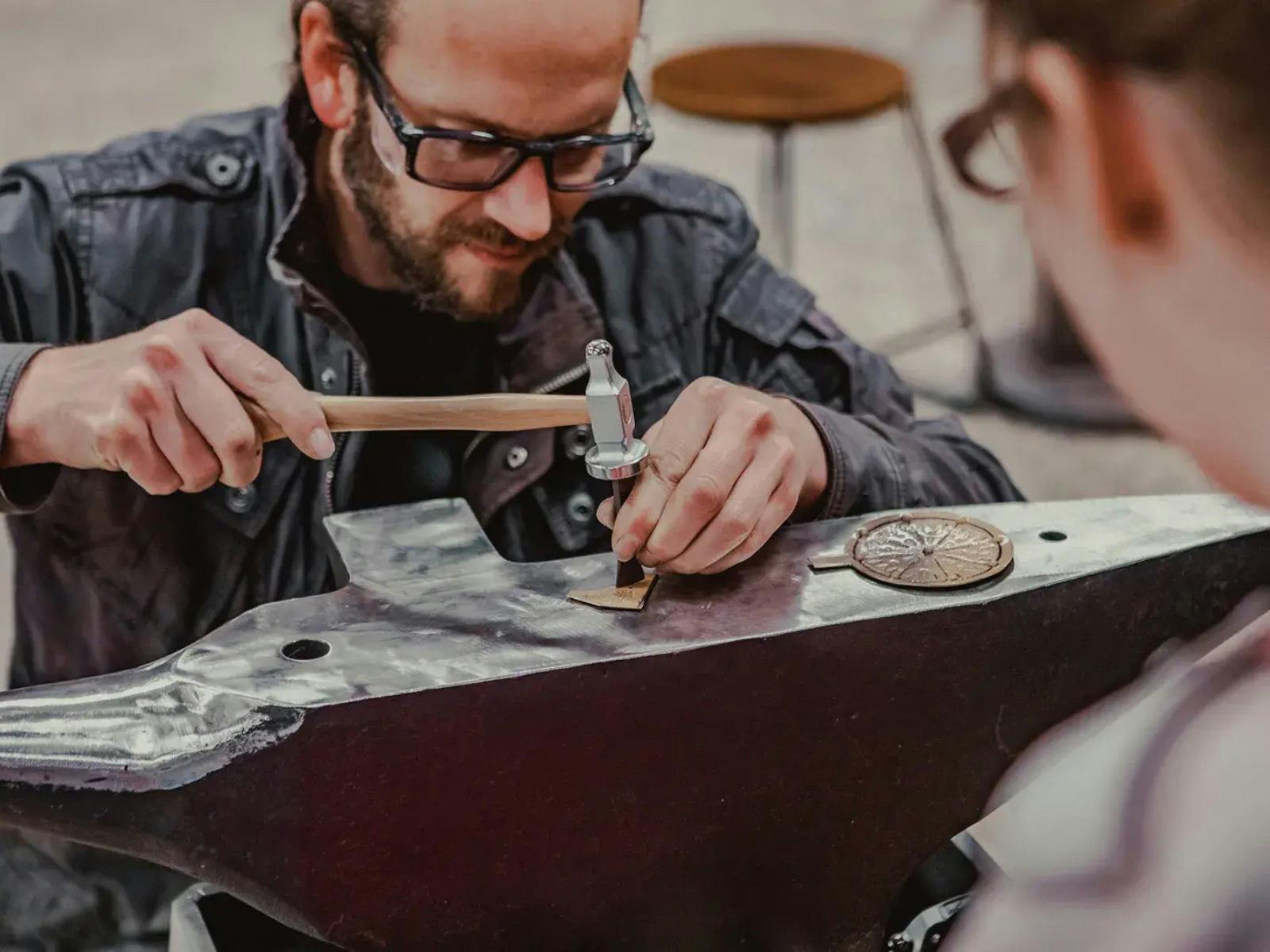 Man hammering metal on anvil while a workshop participant looks on