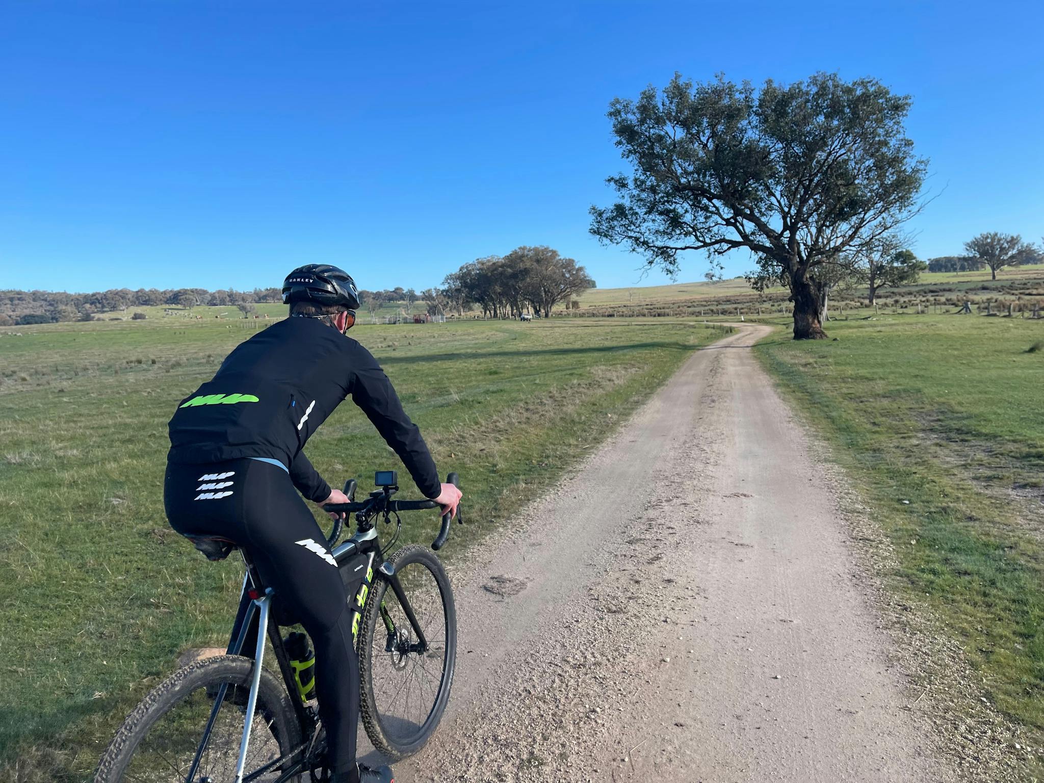 Cyclist in black on gravel road grass on either side of road tree in background and blue sky