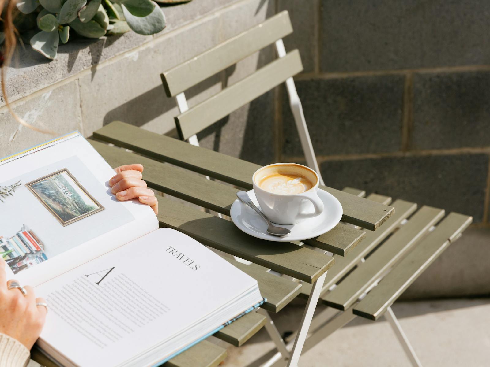 An open book and a partially drunk coffee are drenched in sunshine at an outside table.