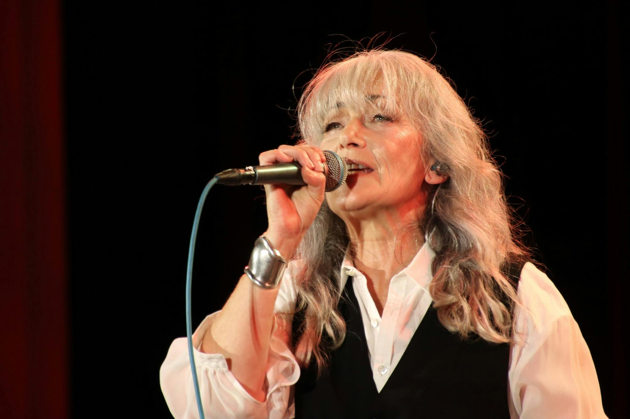 A blonde woman with a black vest and white shirt sings into a microphone with a black background