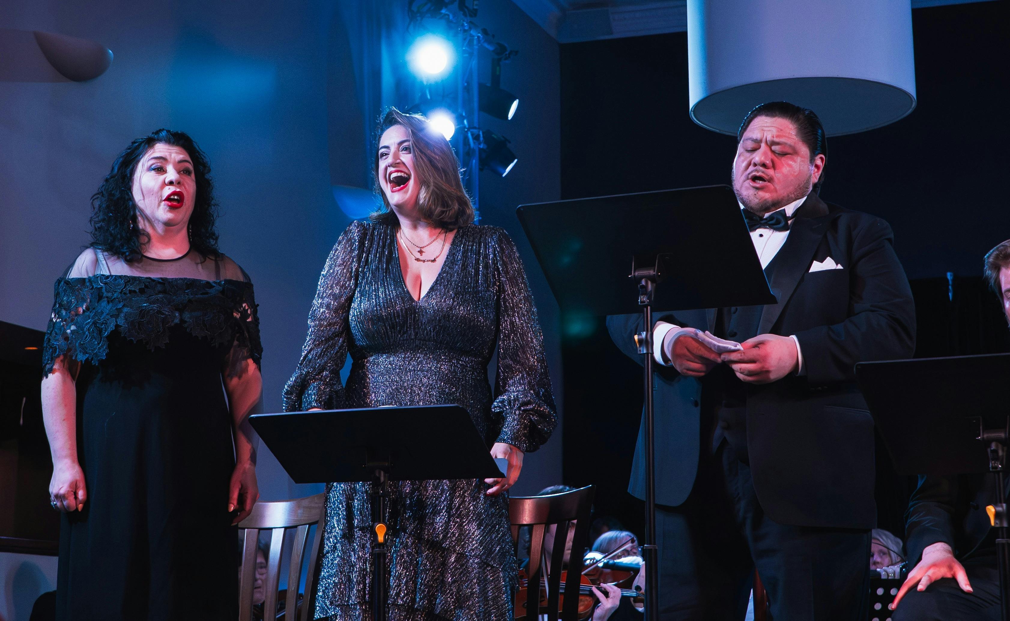Three singers on stage singing opera in front of music stands. 2 females and 1 male.