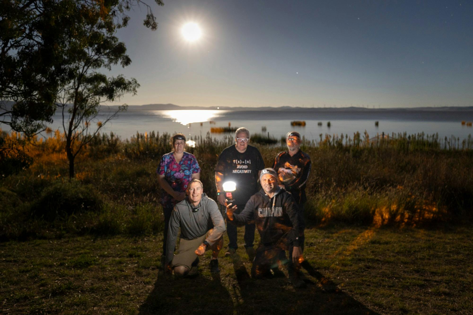 Photographers pose beside a lake under the full moon, with tripods and a camera flash.