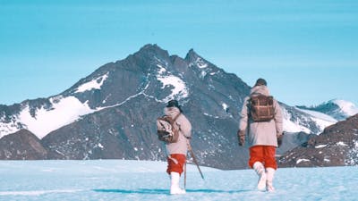 Two hikers walking on snowy ground, in front of snow-covered peaks.