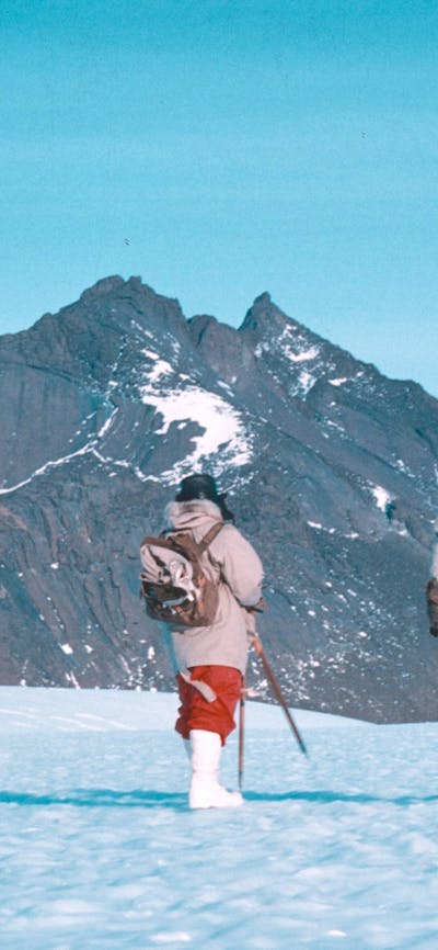 Two hikers walking on snowy ground, in front of snow-covered peaks.