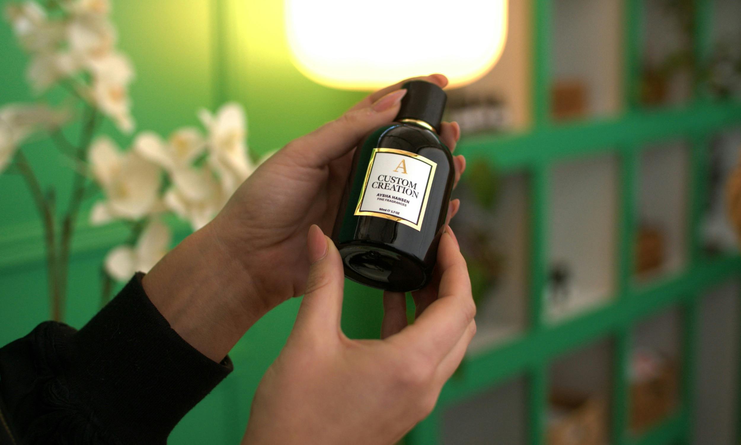Close up of a person holding a black Aysha Hansen Custom Creation perfume bottle against a green bac