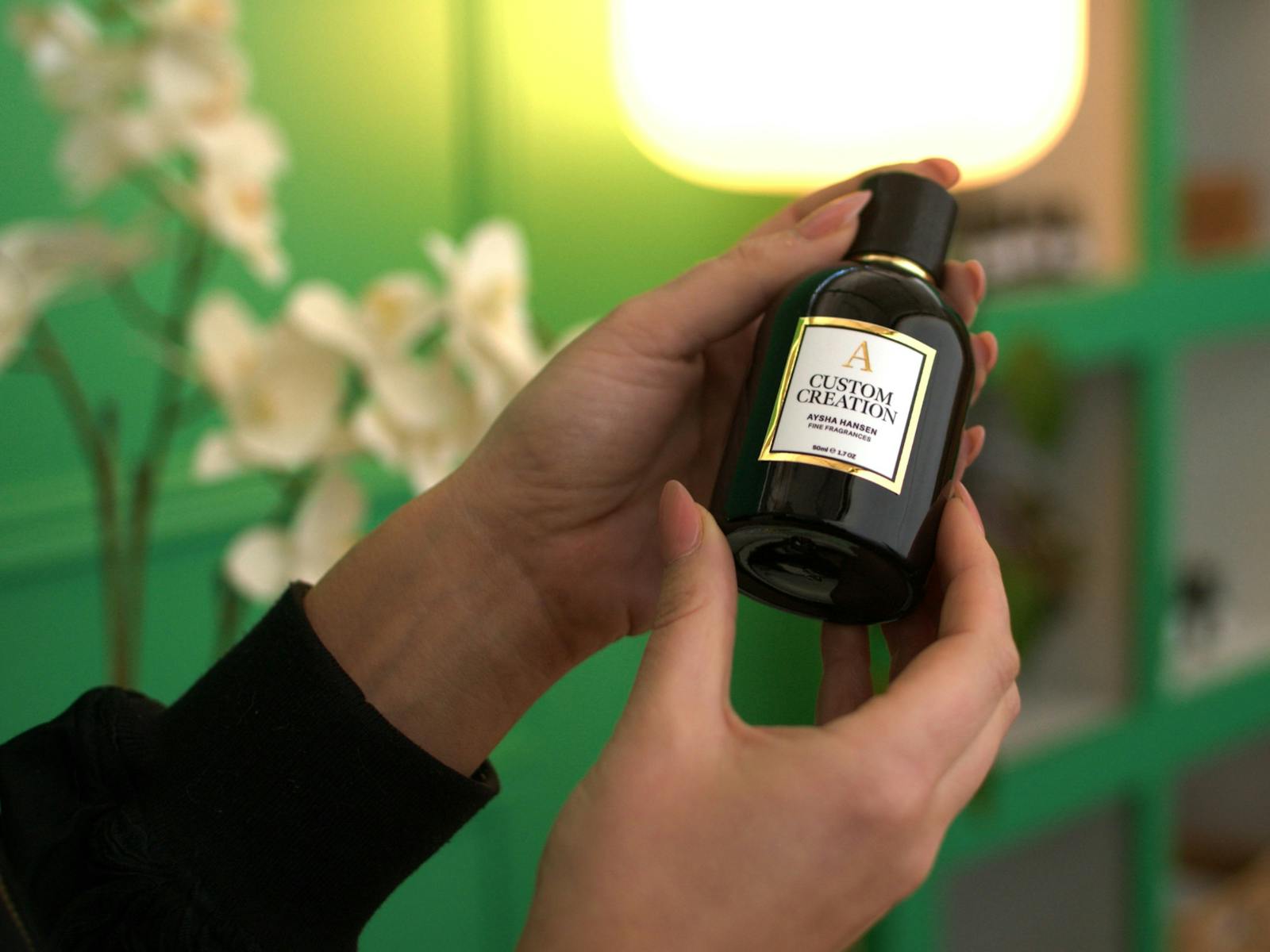 Close up of a person holding a black Aysha Hansen Custom Creation perfume bottle against a green bac