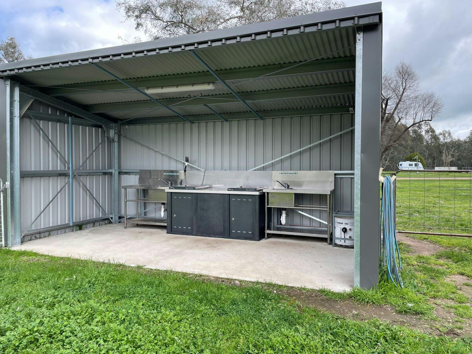 Tin shed looking shelter with BBQ and sink, set on a concrete pad next to green grass