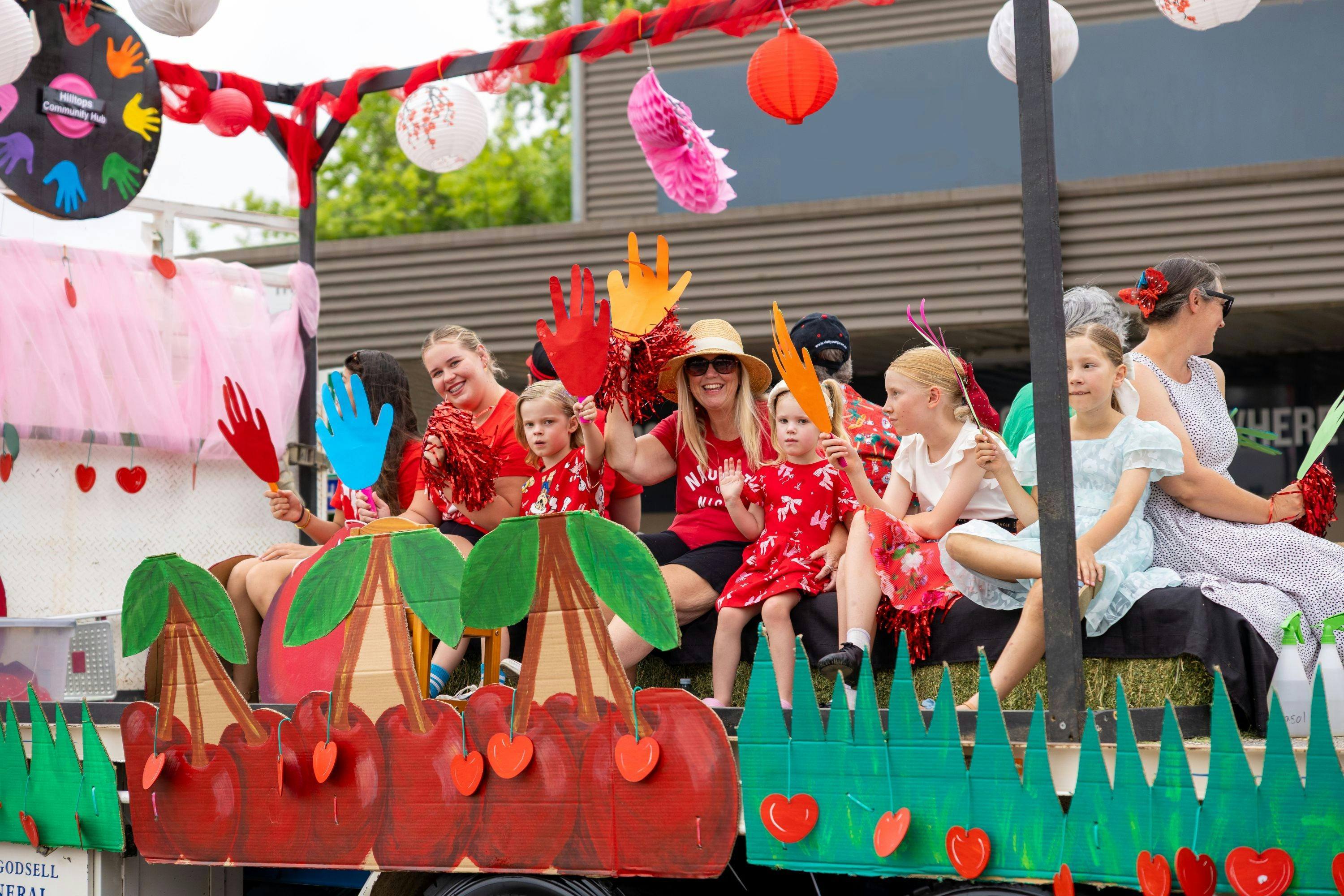A cherry-themed float from the street parade, adults and children waving big paper hands.