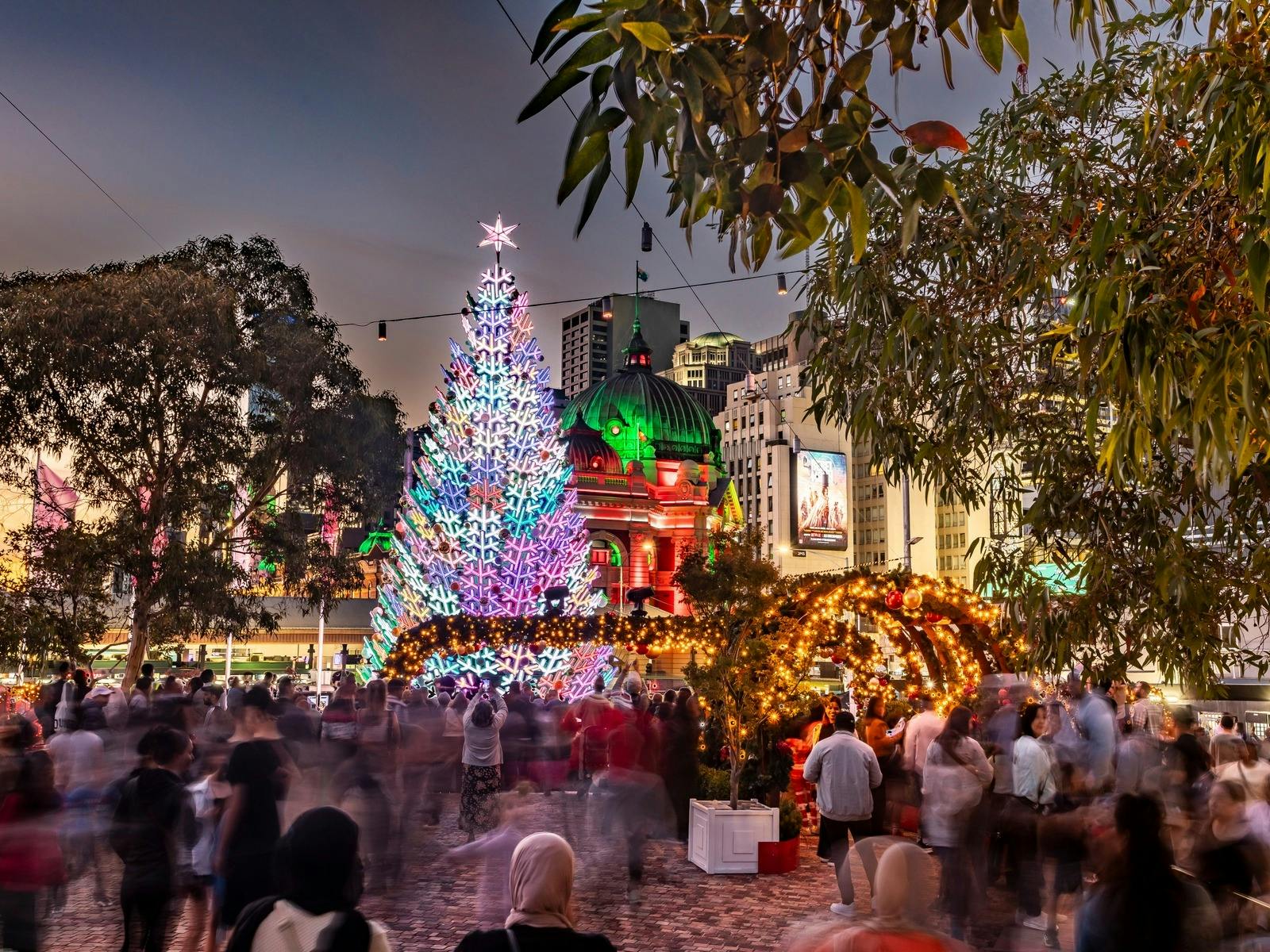 A giant Christmas tree illuminated in rainbow colours at Fed Square. Many people are gathered around