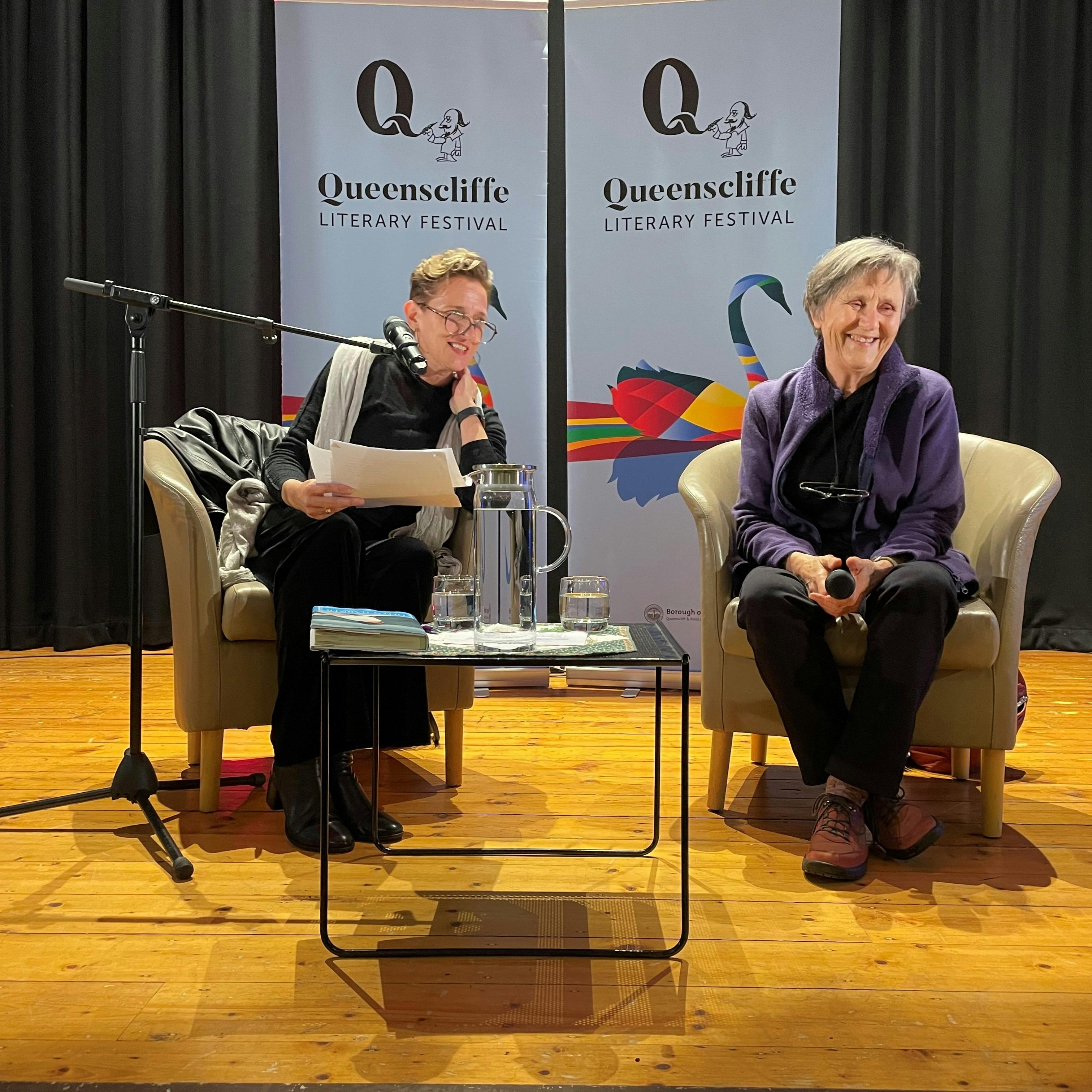 Two women are seated facing the audience, angled to each other. The QLF banners are behind them