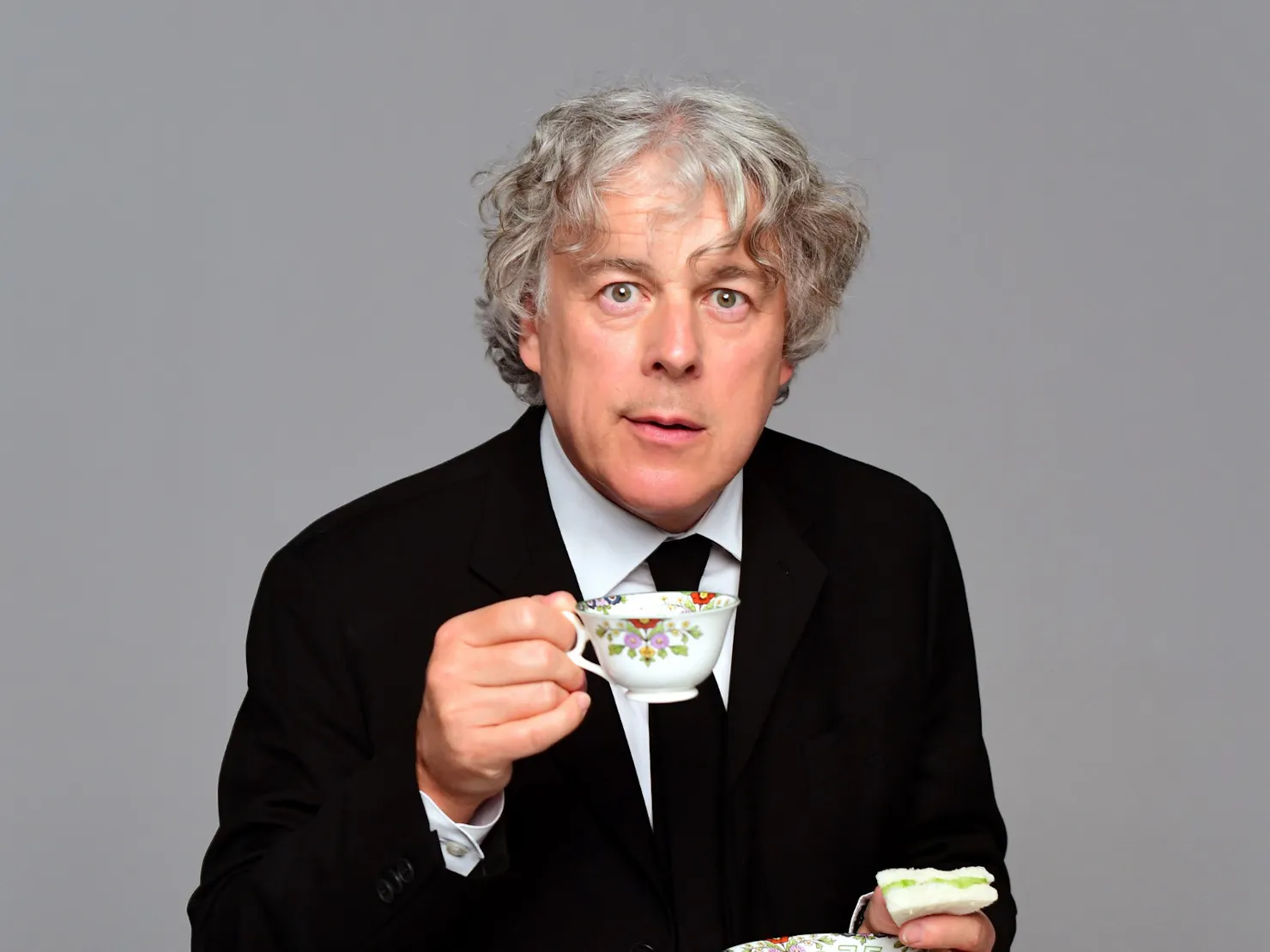 A middle aged man holding a cup of tea and a small sandwich.