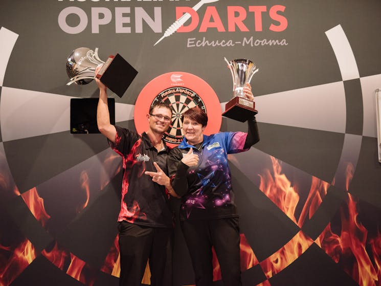 2 people standing in front of a dart board holding trophies