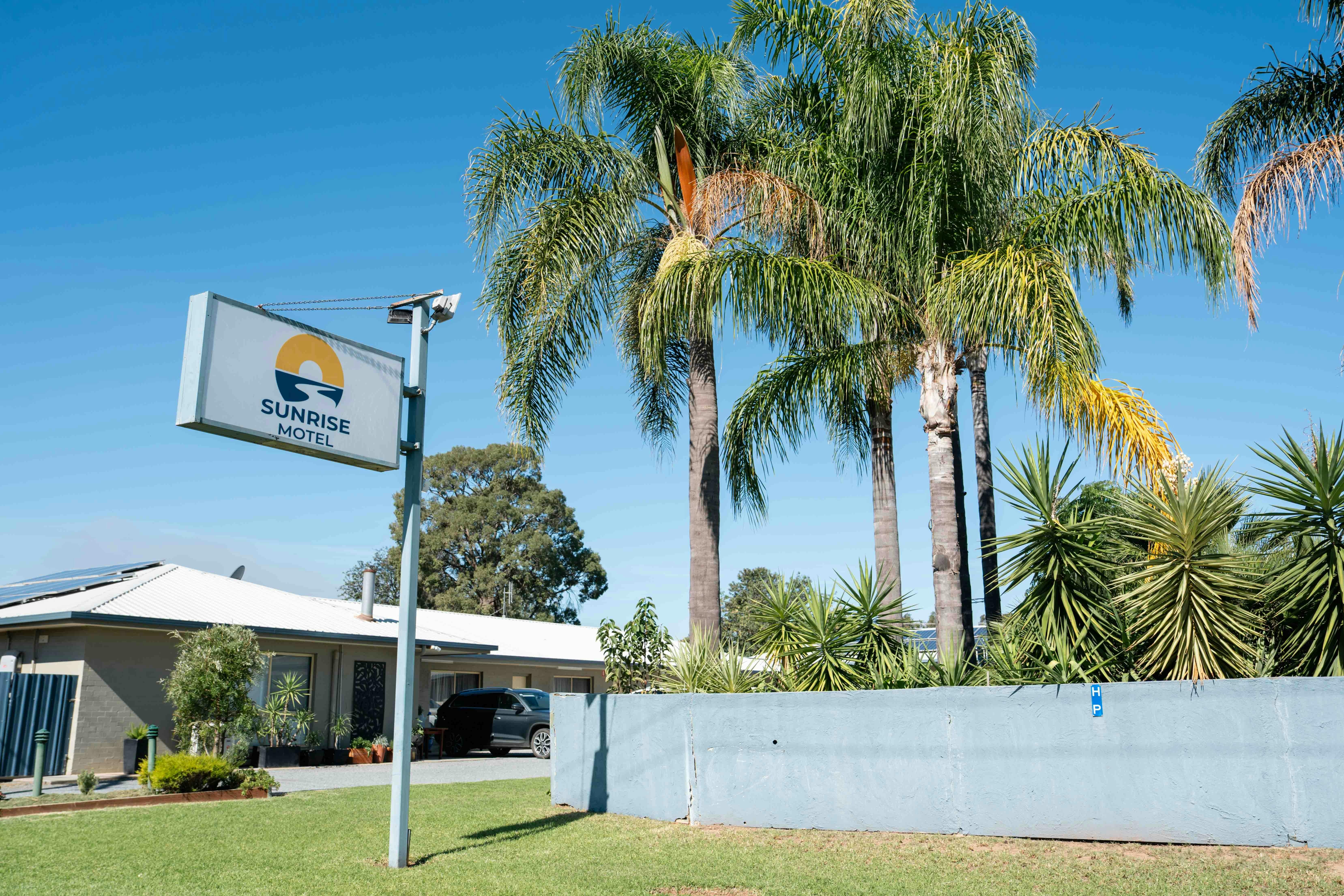 Das Sunrise Motel mit seinem Eingangsschild und der Beschilderung an der Hauptstraße ist von üppigen grünen Palmen umgeben.