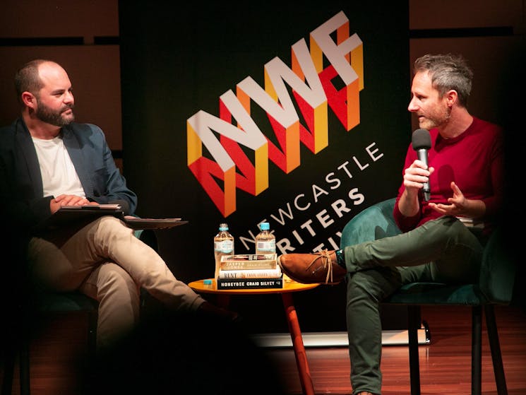ABC Newcastle's Dan Cox seated left speaks to writer Craig Silvey on stage with the festival banner.