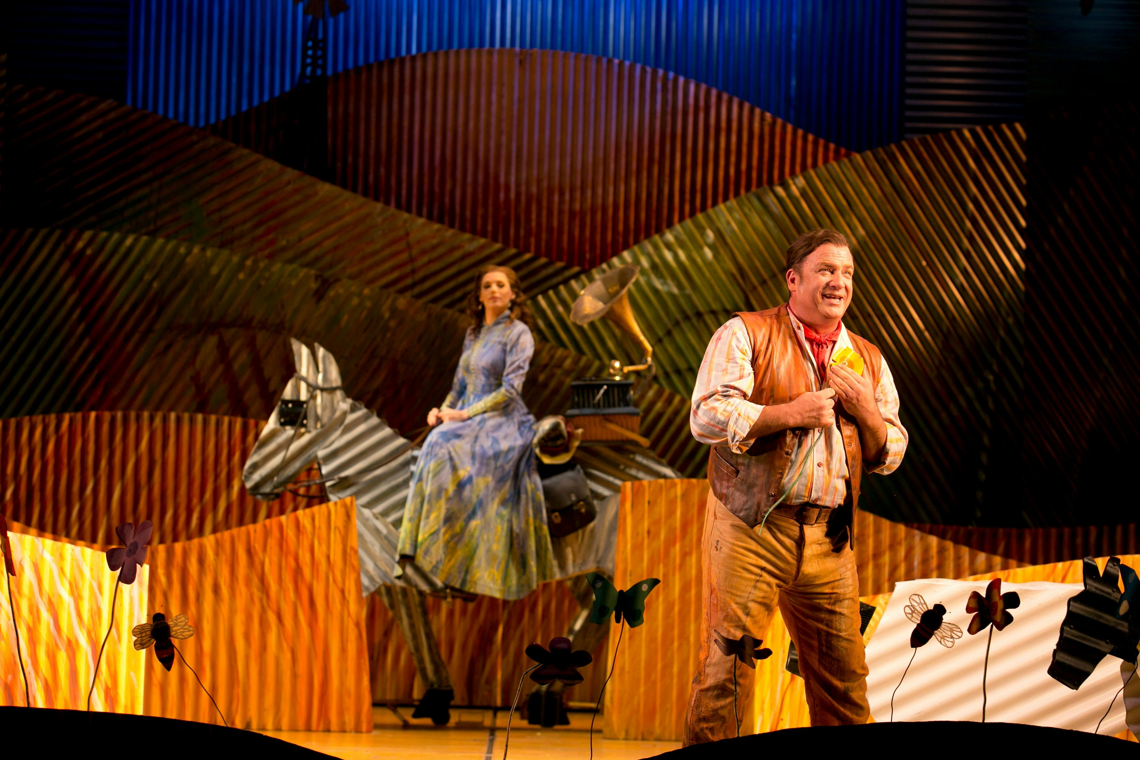 Performers on a colourful stage with a metal horse prop in a whimsical rural setting.