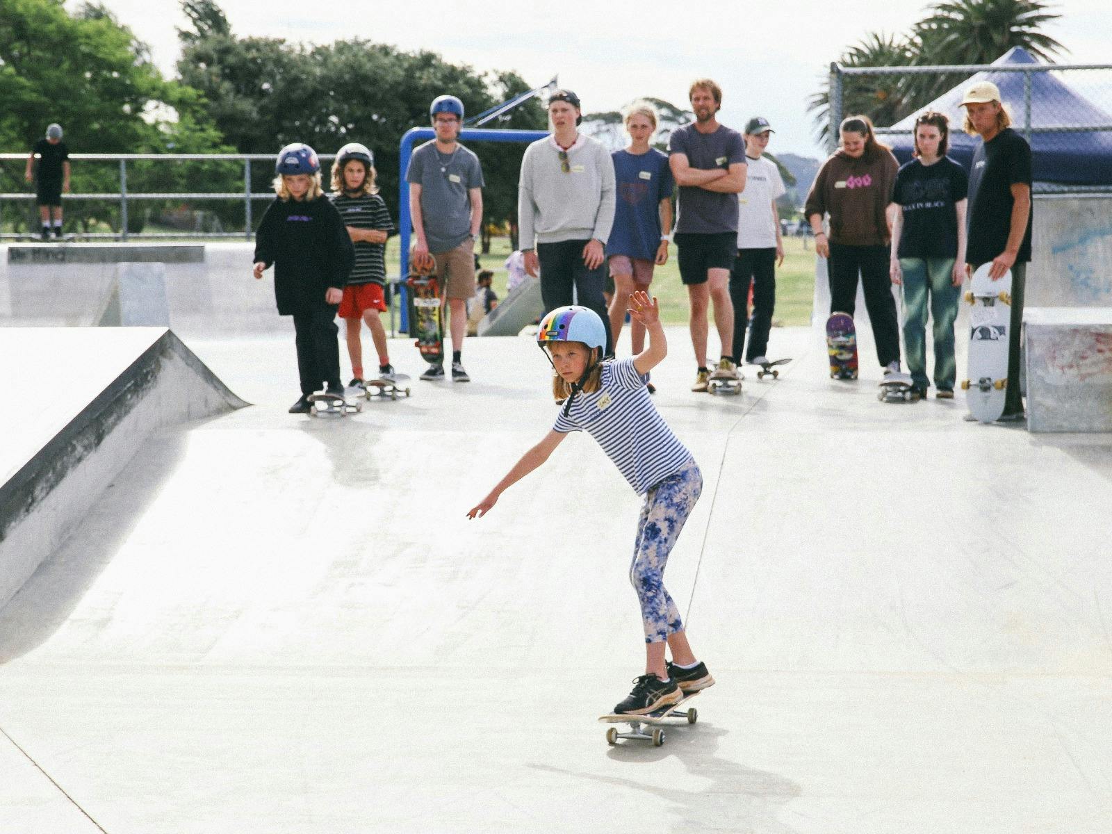 A small crowd looks on from the top of a skate ramp where a young girl student skateboards down