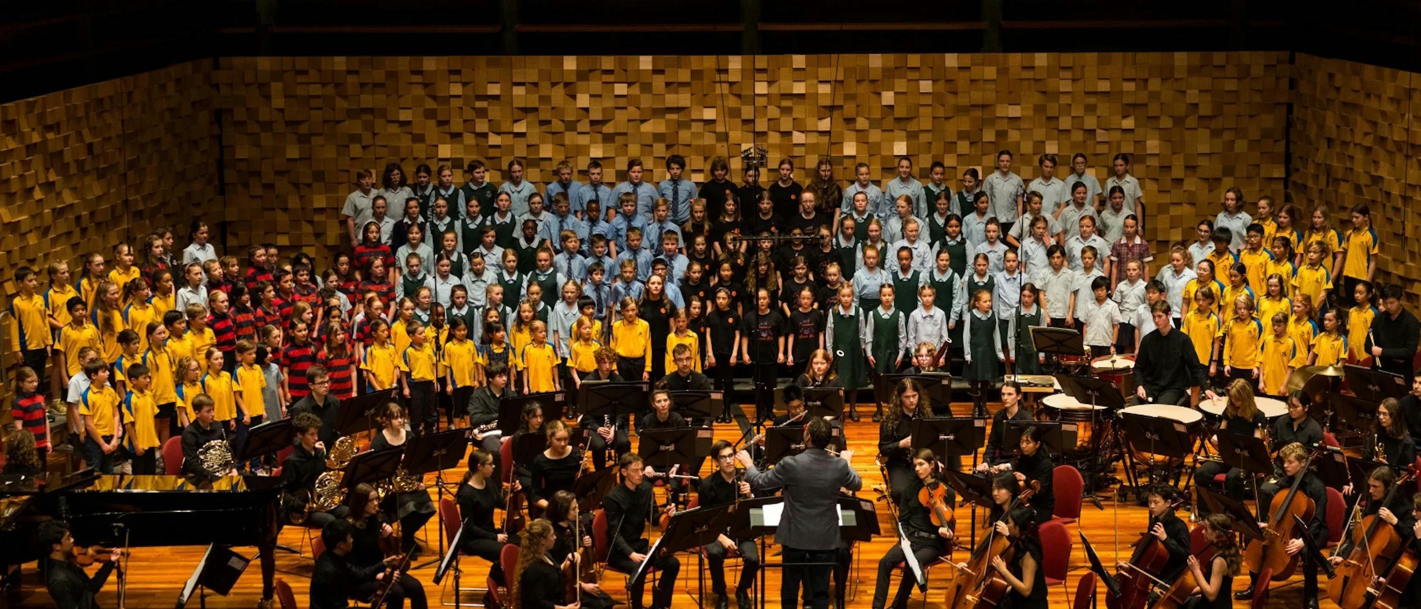 Multiple school choirs stand together on stage with an orchestra playing