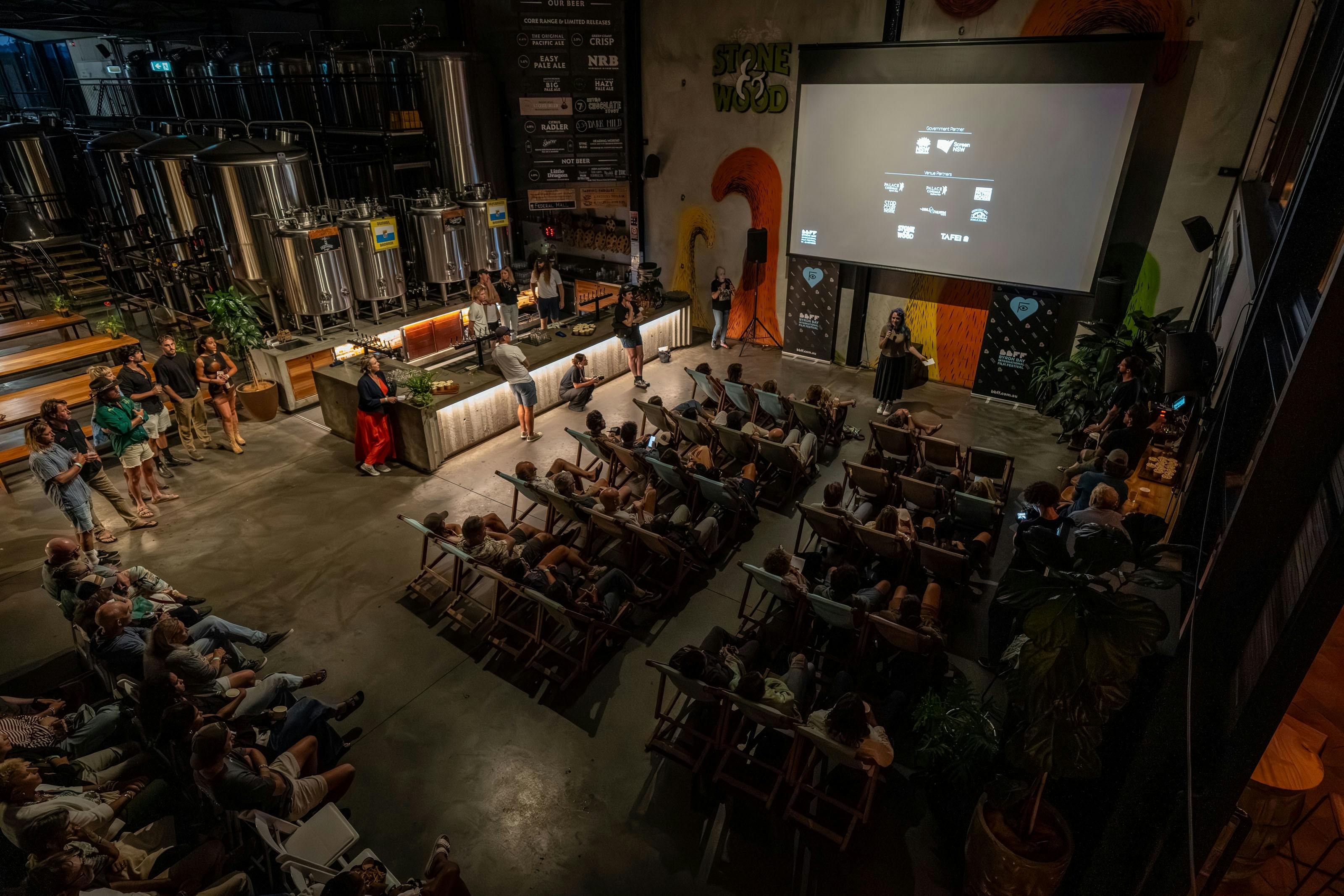 A scene of a crowd of people watching a screen in a beer brewery