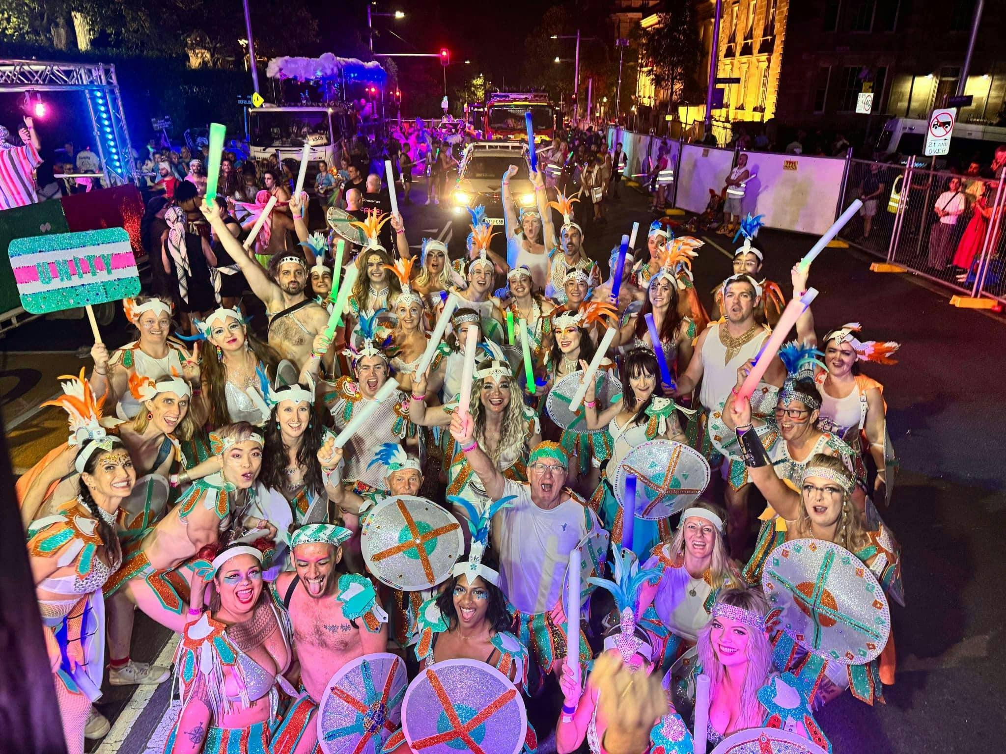 A group of people in rainbow warrior costumes at a mardi gras parade, smiling and cheering.