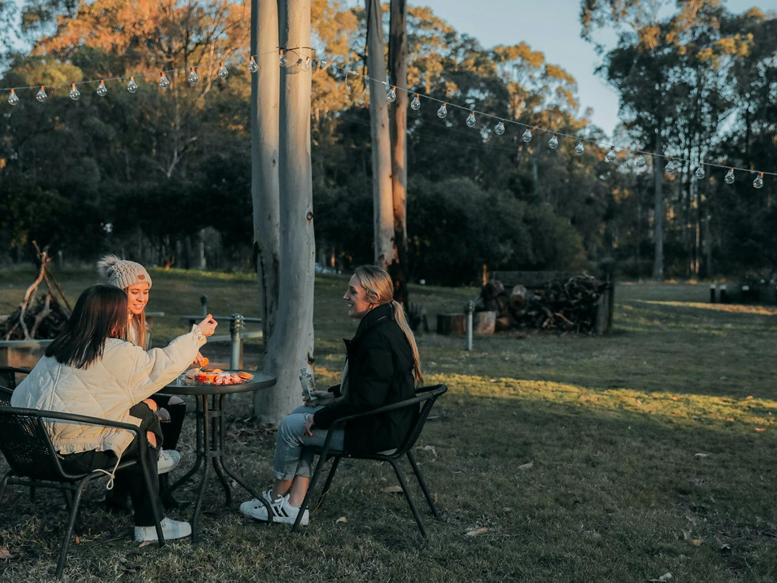 잔디가 우거진 야외 공간에 함께 앉아 자연으로 둘러싸인 치즈 보드를 즐기는 세 명의 여성
