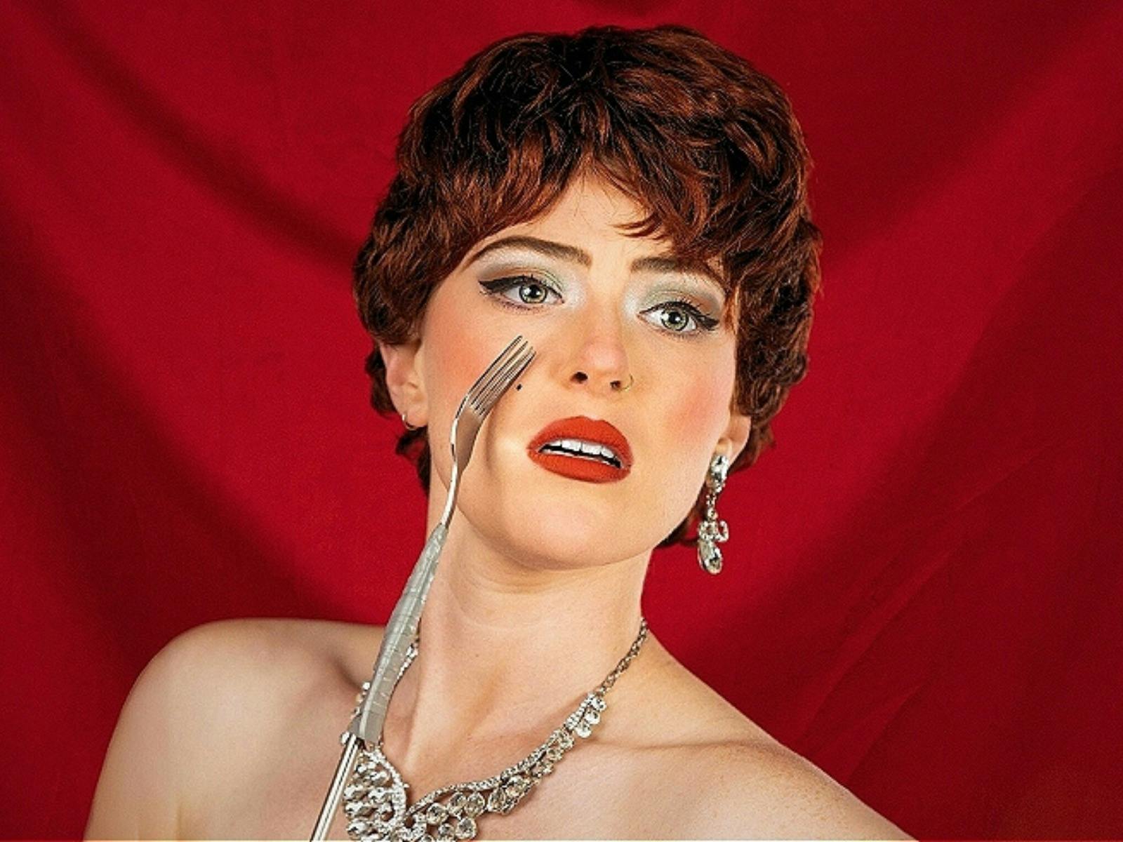 A person with auburn hair, bold red lipstick, and silver jewelry holds a fork against a rich red bac