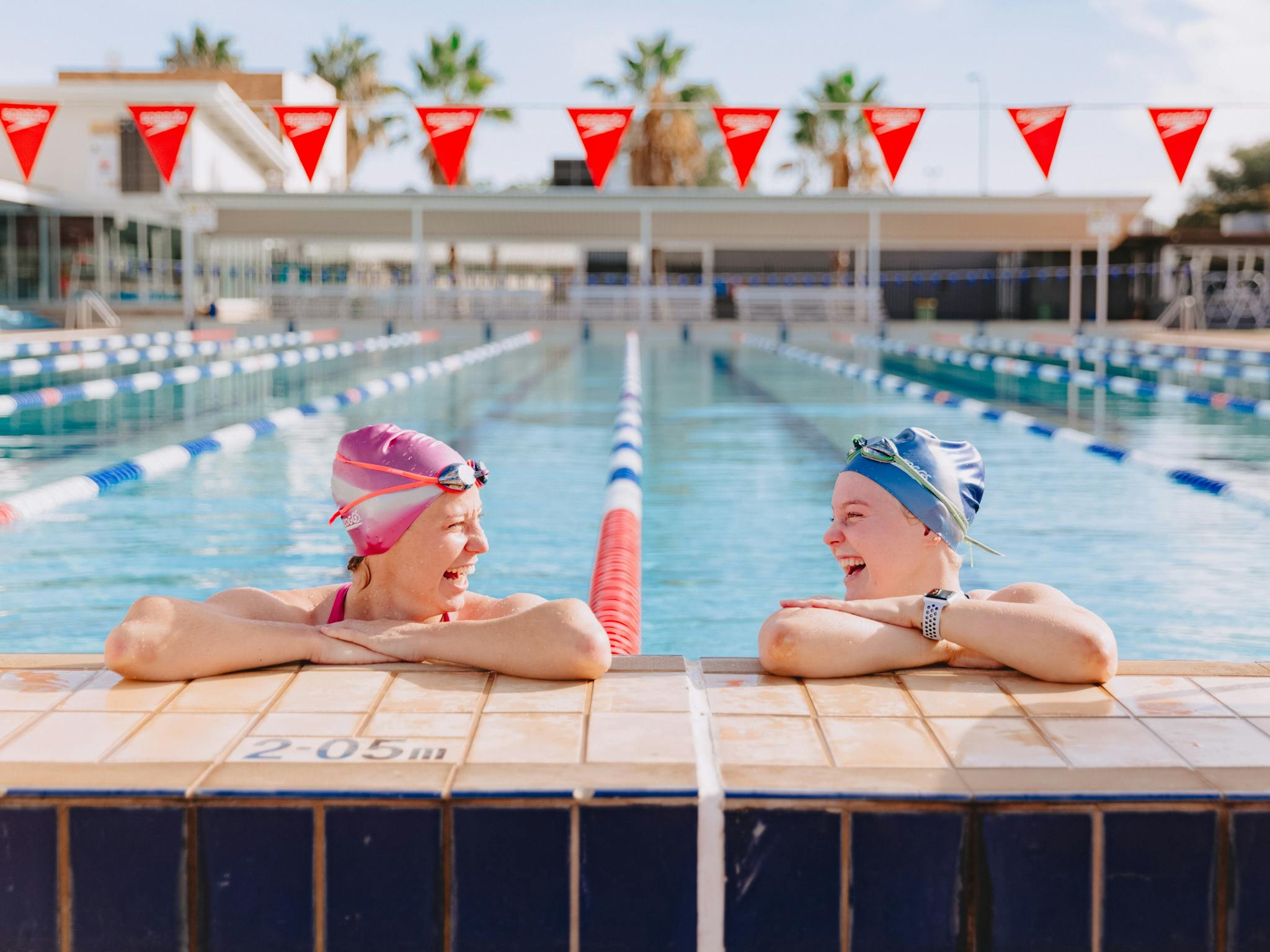 Two swimmers perched up on the edge of the pool smiling at each other