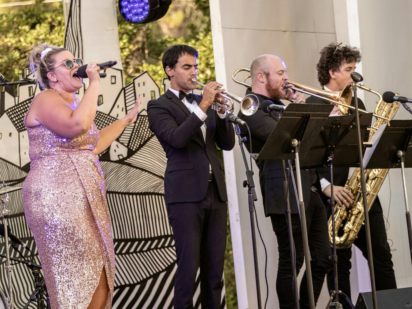 A singer in a sparkly dress and glasses performs with three brass musicians in suits