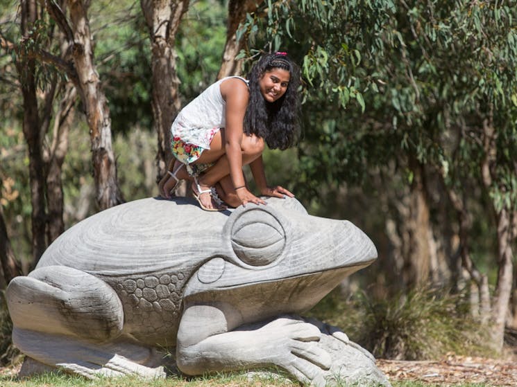 Large sandstone frog sculpure with a young person kneeling on top of it for photo.