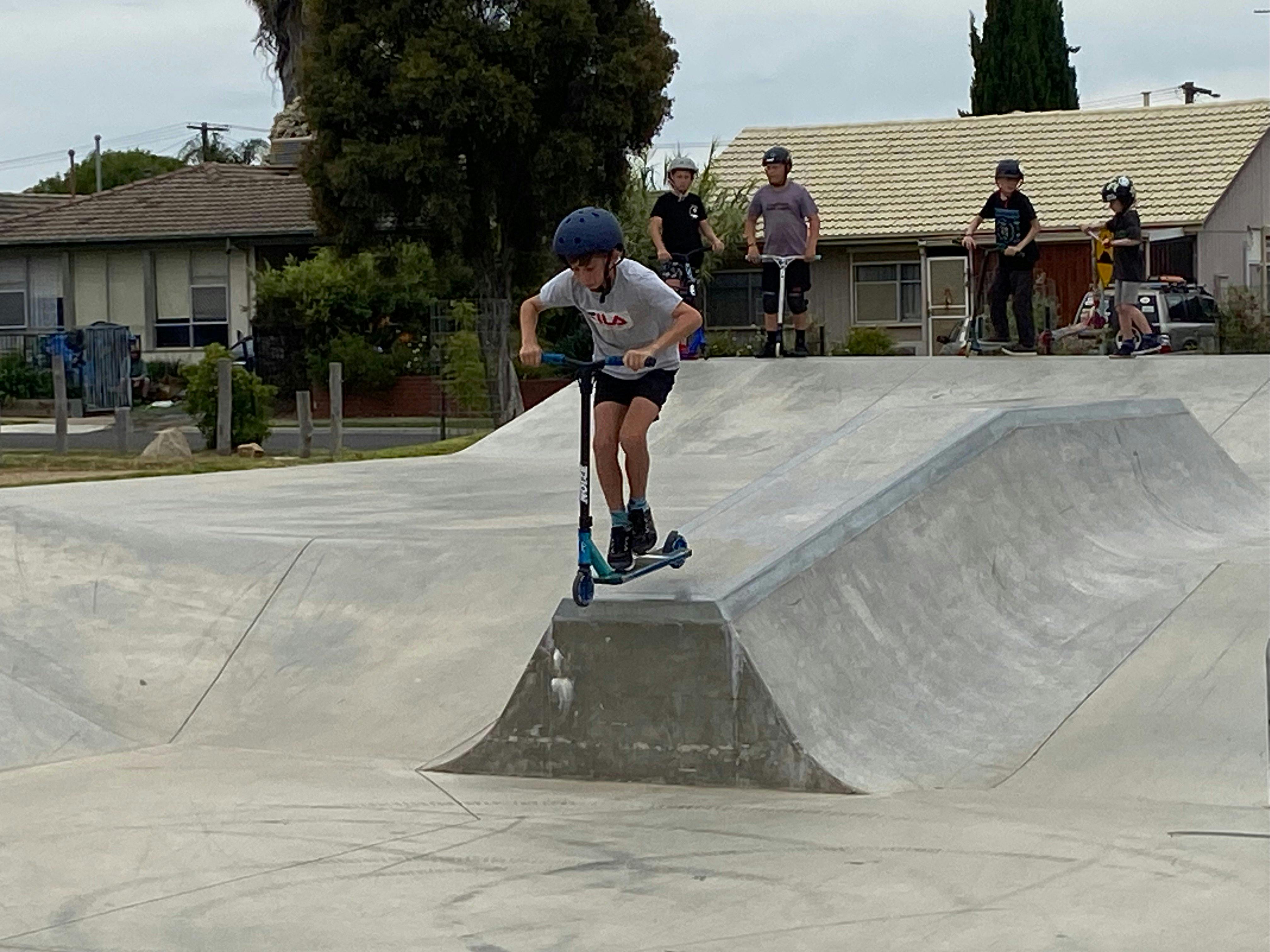 Scooter rider doing trick on cement with several other waiting to go on park houses in background