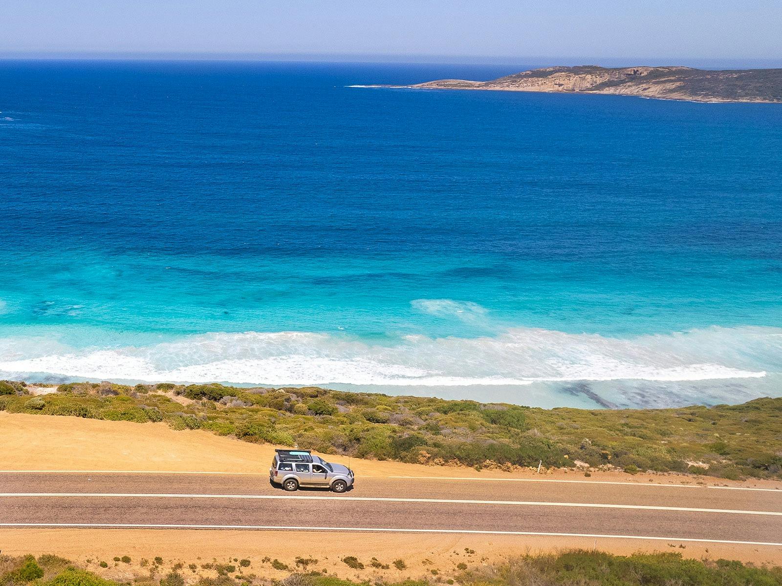 An aerial image of a car driving past coastline with turquoise blue water