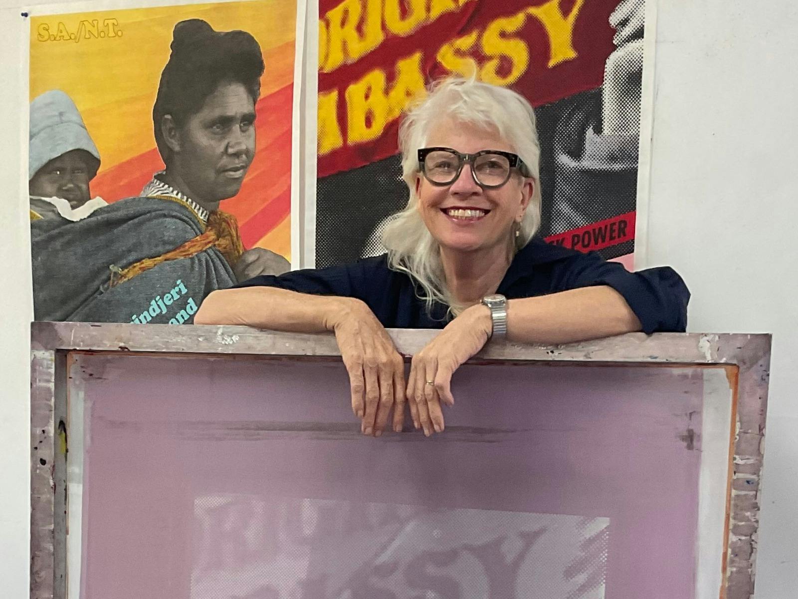 A woman with blonde hair and glasses leaning on a purple artwork piece.